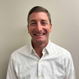 Meet Our Team: A man with short brown hair is smiling at the camera. He is wearing a white button-up shirt and standing against a plain light-colored wall.