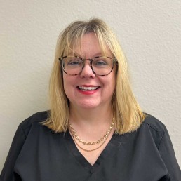 Meet Our Team: A woman with blonde hair and glasses, wearing a black top and gold necklace, is smiling in front of a plain light-colored wall.